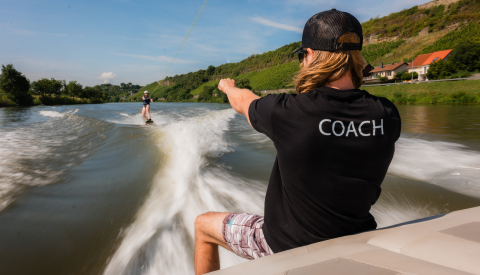 Image d'un animateur de wakeboarding assis sur la partie arrière d'un bâteau en train de donner des instructions à un enfant qui est en train de faire du wakeboard derrière la bâteau